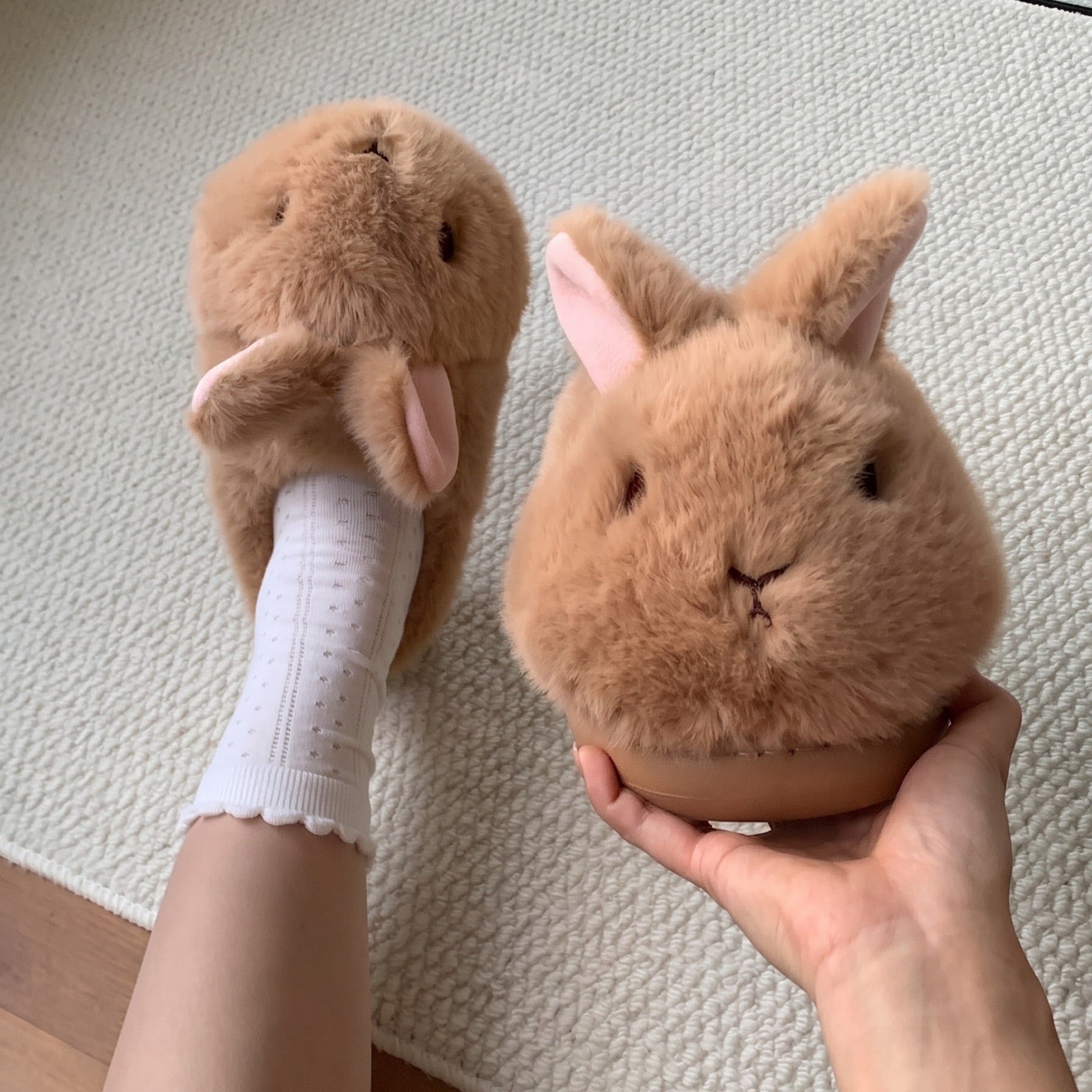 A pair of plush bunny slippers in a warm tan-brown color, with one being worn on a foot in a white sock and the other held in a hand to show detail, set against a textured cream rug.