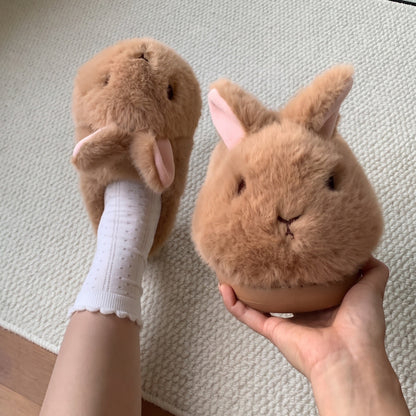 A pair of plush bunny slippers in a warm tan-brown color, with one being worn on a foot in a white sock and the other held in a hand to show detail, set against a textured cream rug.